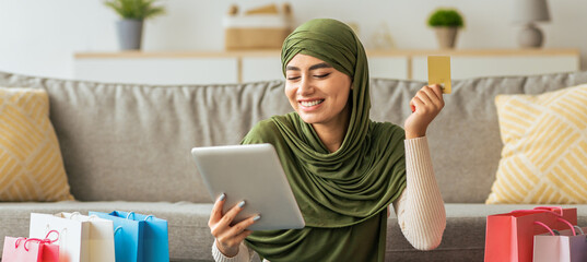 A woman wearing a green headscarf smiles as she holds a tablet and a credit card. She sits in a comfortable living room surrounded by colorful shopping bags, enjoying online shopping.