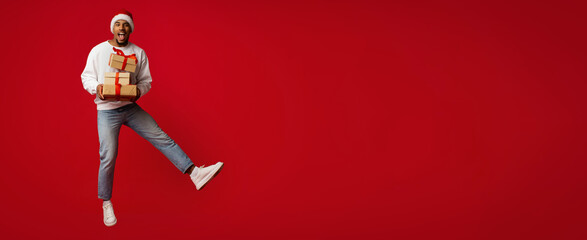 A man wearing a Santa hat joyfully jumps in the air while holding wrapped gifts. The vibrant red background enhances the festive mood. It's a lively celebration of the holiday spirit.
