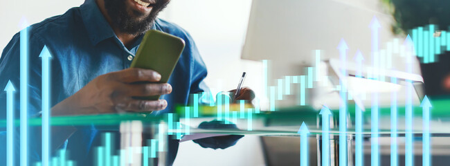 A business professional smiles while using a smartphone in a bright office. He takes notes and reviews market trends displayed through graphics on his screen, focusing intently on his work.