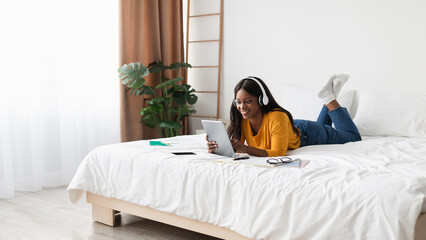 A young woman relaxes on her bed, using a tablet and wearing headphones. She appears focused and engaged while studying in a sunny and inviting room.