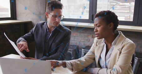 Discussing two colleagues pointing at laptop and holding document in modern office, wearing blazers