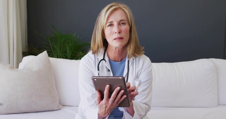 Holding tablet, mature woman in white coat consulting on living room sofa, stethoscope visible