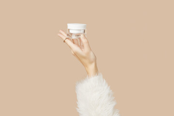 A womans hand in a sweater holds a jar of cream on a beige background. Body care beauty treatment