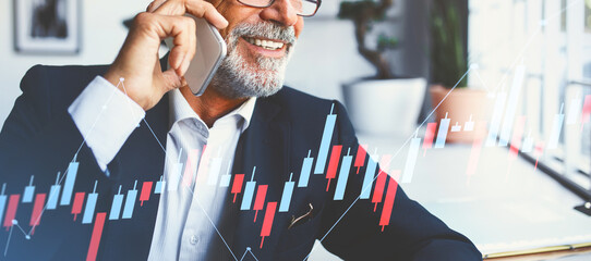 An older businessman in a suit and glasses uses his smartphone to communicate happily while working in a stylish office. He engages in discussions related to business and finance.