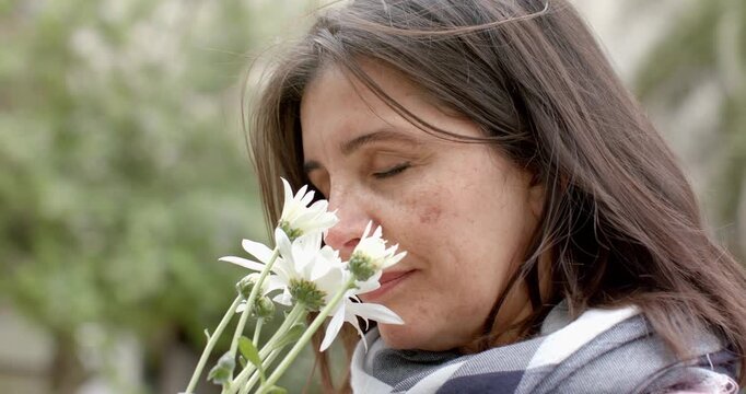 Hispanic woman holding daisies in courtyard, gazing, lifting daisies to nose, smelling and smiling