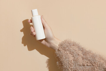 A womans hand in a sweater holds a bottle of cream on a beige background. Body care beauty treatment