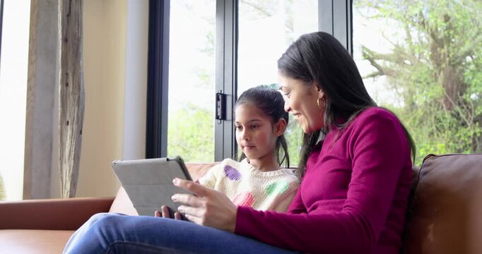 Settling on brown leather sofa, Indian mother guiding daughter tapping tablet for learning at home