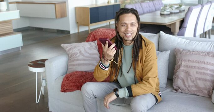 Man in bomber jacket answering call to chat, smiling, speaking on speakerphone on gray sofa