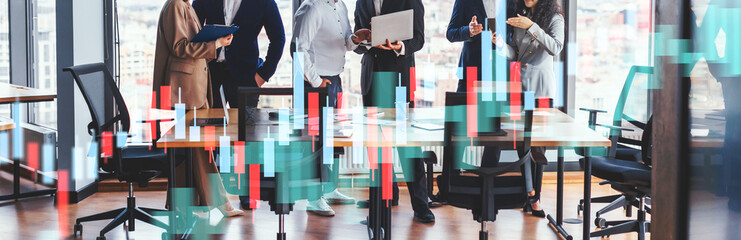 A group of business professionals stands around a large wooden table discussing projects and analyzing data. They use laptops and tablets, sharing ideas in a bright, modern office.