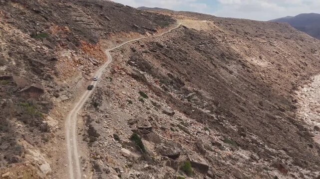 Aerial view of driving cars in dry valley in the island of Socotra. Motion drone video of moving cars on bumpy road surrounded by stones and dessert plants. Following cars from the air. Off-road drive