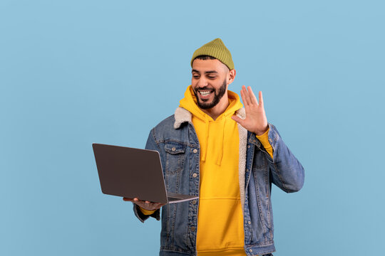 A man wearing a yellow hoodie and denim jacket is smiling and waving at the camera while using his laptop. The bright blue background adds a cheerful vibe to the moment. - Powered by Adobe