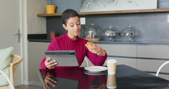 Woman entering kitchen placing tablet on table glancing screen and biting croissant to multitask
