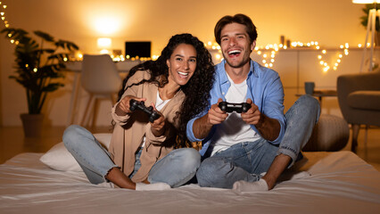 Cheerful couple spends quality time playing video games together in their stylish bedroom at night. They are relaxed and engaged, showcasing a joyful weekend atmosphere.