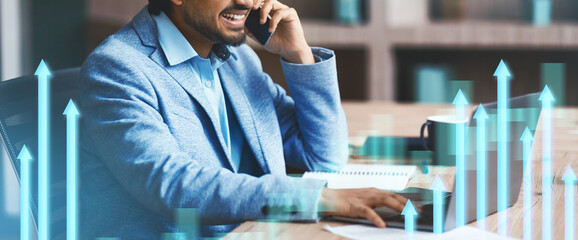An Indian businessman is engaged in a phone conversation while working on his laptop indoors. He demonstrates effective communication skills and a productive work environment at the office.