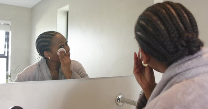 After makeup removal, African American woman pressing cotton pad across face at bathroom mirror