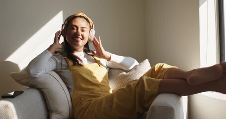 Asian woman on sofa reaching for wireless headphones, placing on ears and relaxing to music - Powered by Adobe