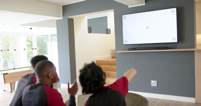 Diverse male friends in jerseys reacting to TV game at home, sipping, flinching at soccer ball