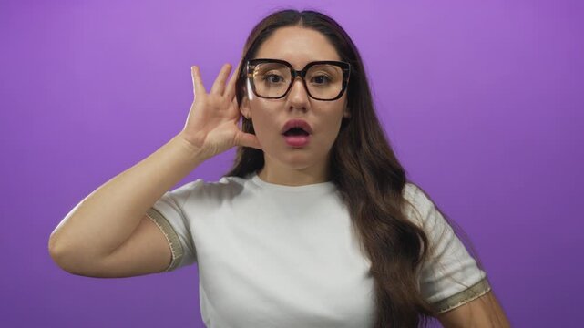 Young hispanic woman wearing white tshirt and eyeglasses cups her ear in purple studio; curiosity focus attention interest.