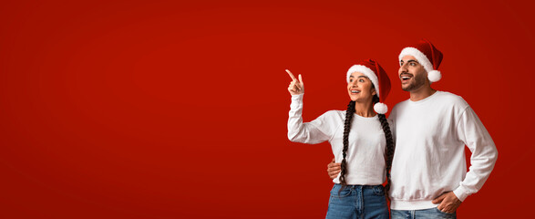 A joyful couple stands together, wearing Santa hats, against a bright red background. They smile...