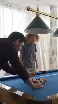 Smiling young man arranging balls in form of equilateral triangle for African American woman playing pool with friends at home during weekend
