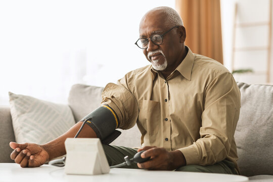 A senior man with glasses is monitoring his blood pressure using a digital device while seated comfortably. Natural light fills the cozy room.
