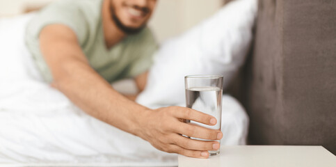 A man is stretching his hand towards a glass of water placed on a bedside table. The cozy bedroom features soft bedding, and he appears refreshed and content.