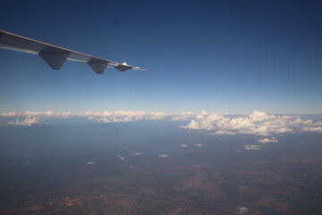 Airplane wing cuts the vast blue sky above fluffy white clouds and land.	
