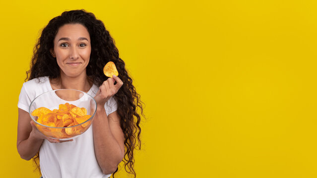 A woman with long, curly hair smiles while holding a bowl of orange chips. She has an engaging expression and stands in front of a bright yellow background, making the scene cheerful and inviting. - Powered by Adobe