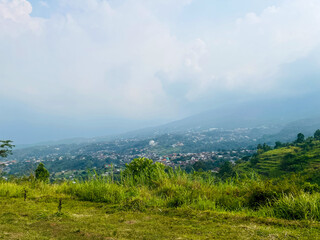 Obraz premium Panorama of rural landscape showing lush green meadows in the foreground. The forest area leads to a valley filled with small buildings. Mountains rise in the distance. The sky is mostly cloudy