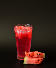 Tall faceted glass of watermelon juice with ice on a black background, pieces of ripe fruit nearby.