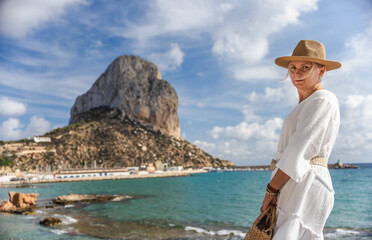 Young woman in a white dress standing by the sea in Calpe, Spain, with the Penon de Ifach rock in the background on a sunny day