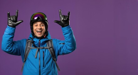 Happy young adult man in blue ski jacket, helmet, and goggles making rock and roll gesture, ready for winter sports adventure on a vibrant purple studio background