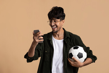 A young man stands smiling and looking at his phone, holding a soccer ball in his other hand. He appears thrilled about recent news or updates, set against a plain background.