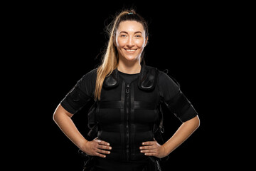 Smiling woman in an EMS fitness suit stands with hands on hips, showcasing electrostimulation training for high intensity workouts and modern gym technology.