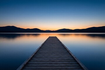 Obraz premium Wooden pier stretching into calm lake water with distant mountains at colorful sunset