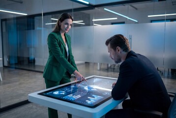 Female advisor showing digital analytics on futuristic touch table to businessman in glass office
