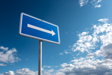 Blue directional sign with a white arrow pointing right, set against a bright, cloudy blue sky. Traffic guidance.