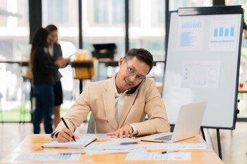 The young man enjoys his work, talking on the phone while using his laptop, communicating business and multitasking, with colleagues behind him.