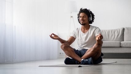 A relaxed black man sits in a lotus position on a yoga mat at home, wearing wireless headphones. He smiles with his eyes closed, embodying tranquility and peace during meditation.