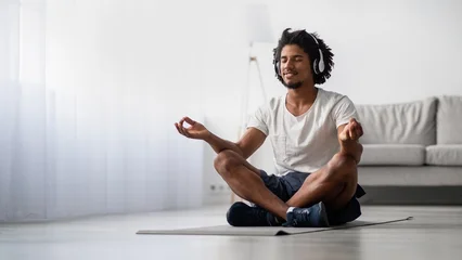 Fotobehang Lotusbloem A relaxed black man sits in a lotus position on a yoga mat at home, wearing wireless headphones. He smiles with his eyes closed, embodying tranquility and peace during meditation.  © Prostock-studio