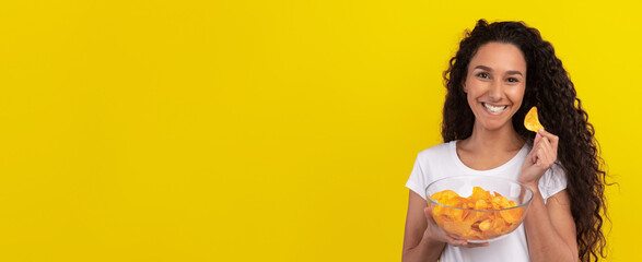 A smiling young woman holds a clear bowl of orange snacks while munching on one. She stands in...