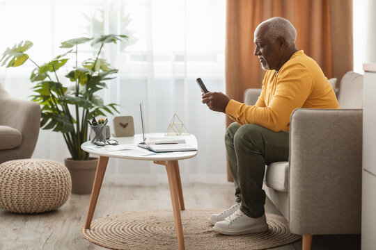 A senior man wearing a yellow sweater sits on a couch, focused on his smartphone. The room has plants, a laptop, and a soft area rug, creating a warm atmosphere. - Powered by Adobe