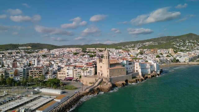 Aerial 4K orbit of San Bartolome church in Sitges, historic cathedral on the Mediterranean coast with blue sea and waves, Catalonia, Spain, drone shot