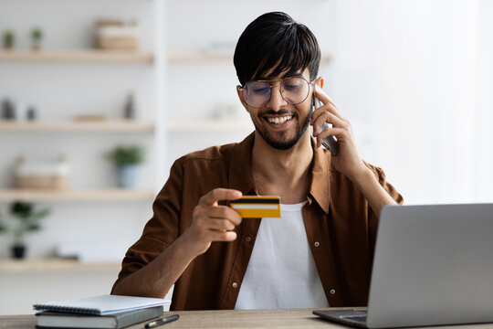 A young man is seated at a wooden desk, smiling as he talks on the phone. He holds a yellow and black credit card in his hand while working on his laptop, creating a busy but cheerful atmosphere. - Powered by Adobe