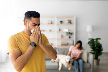 A young man is sneezing and wiping his nose with a tissue in a modern living room. Nearby, a woman...