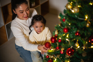 mother and toddler girl decorating Christmas tree in home at night