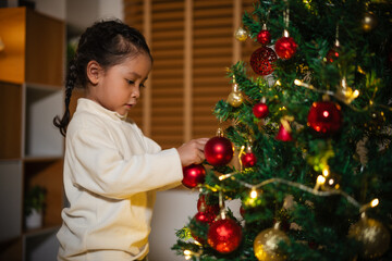 toddler girl decorating toy ball on Christmas tree in home at night