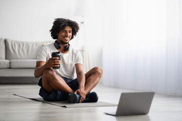 Sporty African American man sits on a yoga mat at home, watching workout videos on his laptop. He holds a fitness shaker and enjoys the guidance of online tutorials.