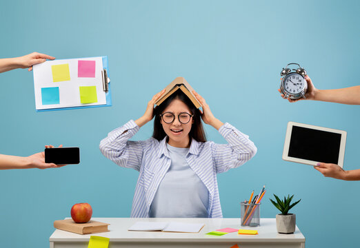 A student appears stressed while juggling various tasks at a busy desk. She holds a book over her head with sticky notes, a clock, a tablet, and a phone presented by hands around her.