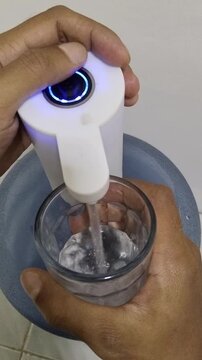 hand filling a glass with clean water using a white portable electric gallon pump. Practical and hygienic solution for everyday drinking water